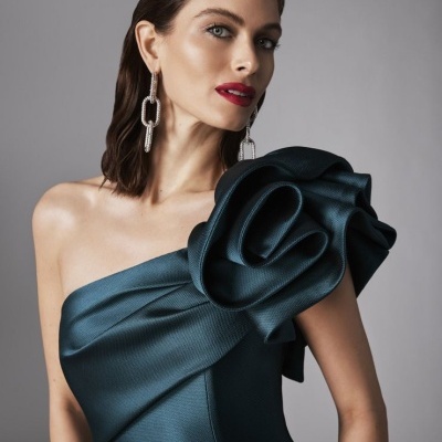 Vestido azul escuro com detalhe em flor num ombro, brincos prateados, fundo cinza