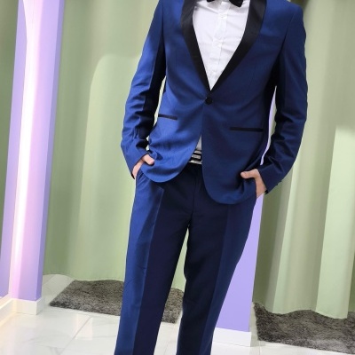 Homem vestido com smoking azul e gravata borboleta preta em sala com parede roxa e cortinas verdes