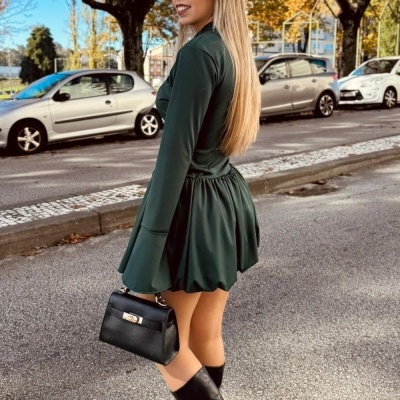 Mulher de vestido verde escuro e botas pretas numa rua com árvores e carros