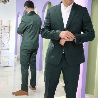 Homem com fato verde escuro, camisa branca e sapatos castanhos em ambiente lilás com espelho e flores.