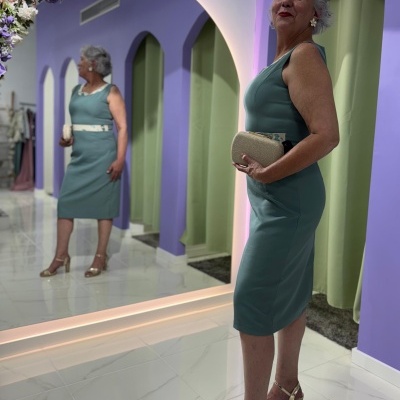 Mulher com vestido verde-azulado e mala bege em interior com espelho e flores