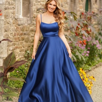 Mulher com vestido longo azul acetinado perto de parede de pedra e plantas.