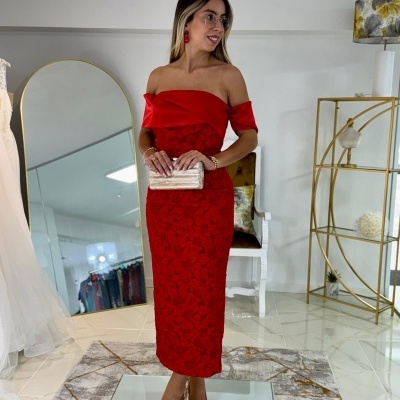 Mulher com vestido vermelho rendado com padrão floral, sapatos nude e bolsa creme em ambiente interior moderno