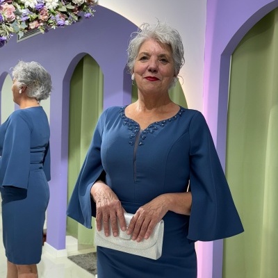 Mulher com vestido azul escuro de mangas largas segurando carteira branca num ambiente com paredes lilás e verde claro