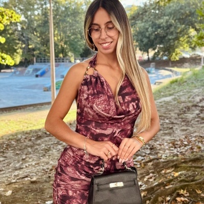 Mulher com vestido curto estampado tons castanhos e roxos, mala de mão preta e acessórios, ao ar livre com árvores ao fundo.
