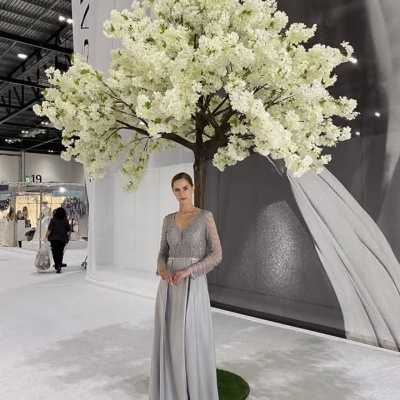 Mulher com vestido cinzento longo ao pé de uma árvore decorativa branca