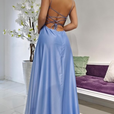 Mulher de costas com vestido azul acetinado de alças finas cruzadas, em interior com flores e sofá roxo.