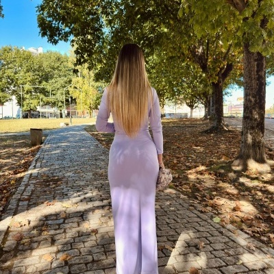 mulher vestida com vestido lilás comprido a caminhar numa calçada de pedra sob árvores