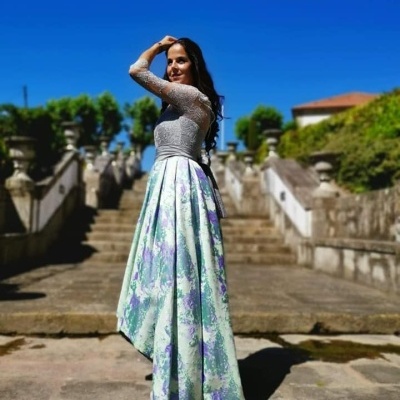 Mulher a usar vestido longo com parte superior prata brilhante e saia florida verde, lilás e branco ao sol em escadaria de pedra.