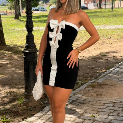 Mulher com vestido preto e branco com laços, sapatos brancos e mala, em parque com árvores e calçada de pedra.