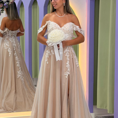 Mulher a usar vestido de noiva bege com renda branca, segurando bouquet branco, num ambiente com espelho e flores decorativas.