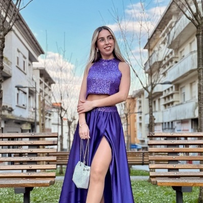 Mulher com vestido roxo de duas peças e mala branca em ambiente urbano com bancos de madeira.