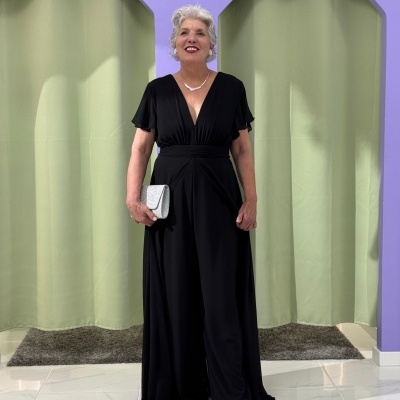 Mulher sénior vestindo macacão preto e segurando clutch cinza prata em ambiente com cortinas verdes e parede lilás.
