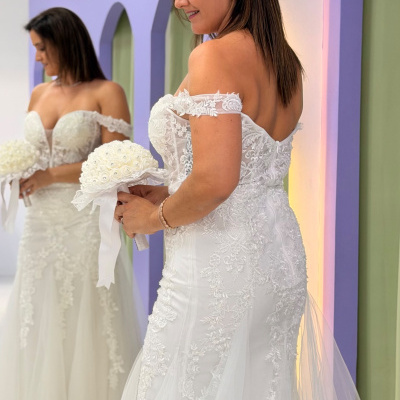 Mulher com vestido de noiva branco rendado segurando ramo branco em frente a espelho
