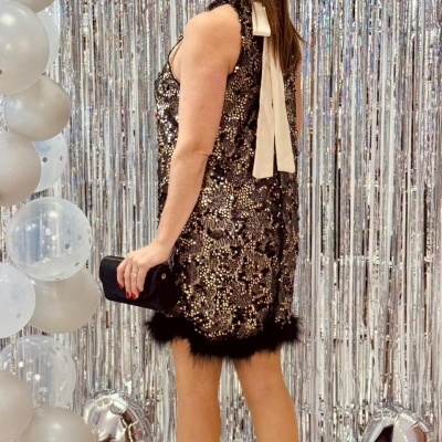 Mulher de vestido de lantejoulas douradas com pena preta e fita branca, sapatos pretos de salto, segurando mala preta, com fundo de cortina prateada e balões prata e brancos