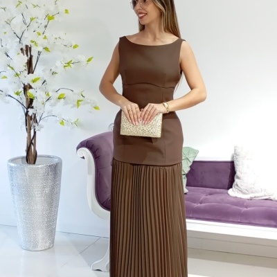 Mulher com vestido castanho e clutch dourada em interior com sofá roxo e vaso com flores