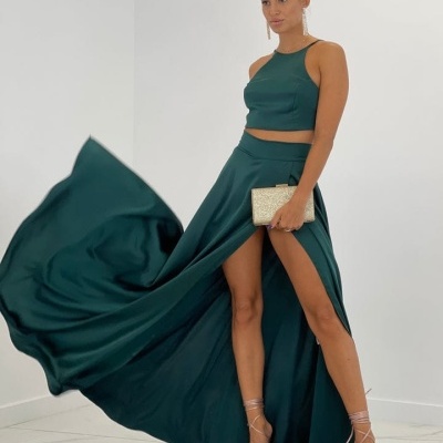 Mulher com conjunto verde escuro de top e saia longa com fenda, clutch dourada e salto alto nude, em ambiente interior branco