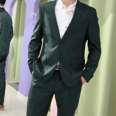 Homem com fato verde-escuro e camisa branca em interior com parede lilás e cortina verde