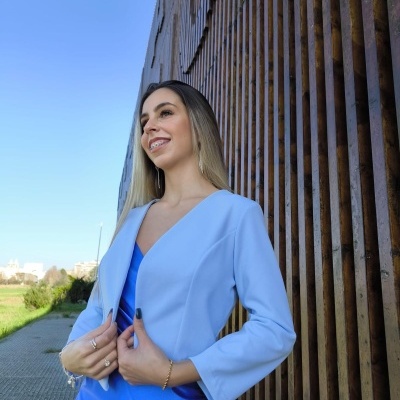 Mulher com casaco azul claro e vestido azul brilhante ao ar livre junto a parede de madeira