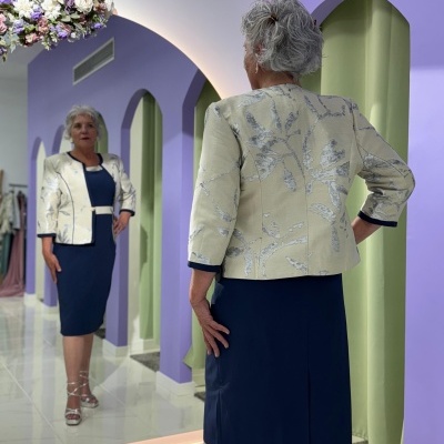 Mulher mais velha veste vestido azul e casaco bege com padrão floral, de pé diante de espelho com paredes lilás