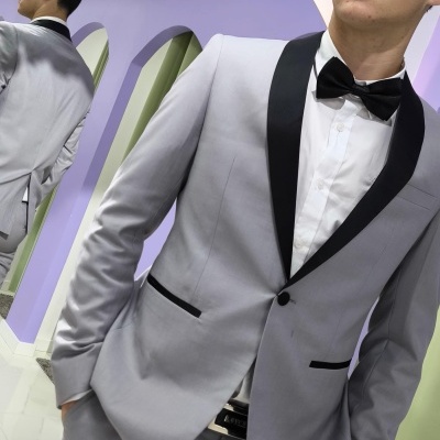 Homem vestido com fato cinzento claro e gravata borboleta preta em frente a um espelho com decoração floral