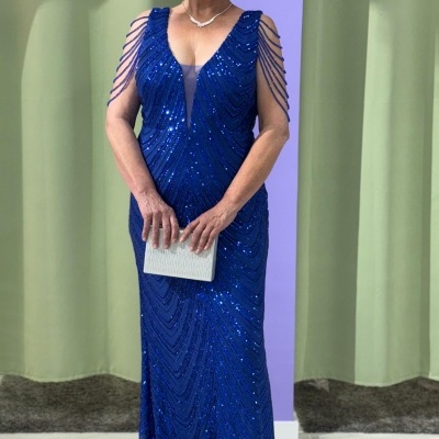 Mulher com vestido azul brilhante e clutch branca em ambiente com cortinas verdes.