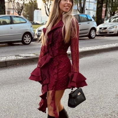 Mulher com vestido vinho bordado e folhos, botas pretas e mala preta na rua com carros e árvores