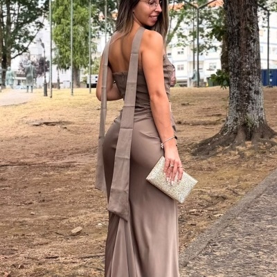 Mulher com vestido castanho longo e alças largas segurando carteira prateada em parque com árvores