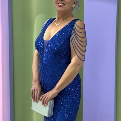 Mulher com vestido azul com lantejoulas e franjas, colar e brincos prateados, segurando carteira prateada, fundo colorido com arco