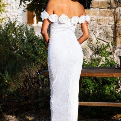 Vestido comprido branco com flores no ombro usado por mulher num jardim com vegetação e pedras