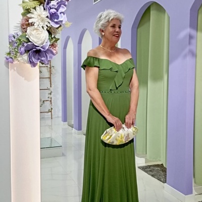 Mulher com vestido verde longo e bolsa branca e amarela em ambiente com parede lilás e arco de flores.