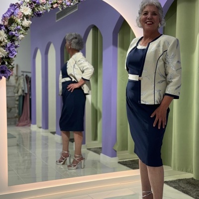 Mulher com vestido azul escuro e casaco bege com padrão floral junto a espelho com decoração floral roxa.