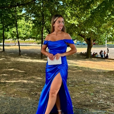 Mulher com vestido azul longo acetinado e racha lateral segurando carteira branca, ao ar livre