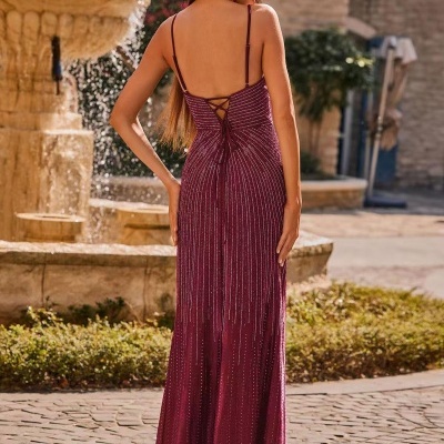 Mulher de costas usando vestido longo vinho com brilho e alças finas numa rua com fonte de pedra ao fundo