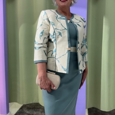 Conjunto feminino vestido azul-petróleo e casaco creme com padrão azul em ambiente interior.