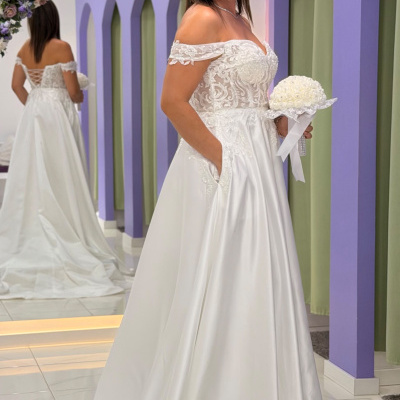 Mulher com vestido de noiva branco com detalhes em renda, segurando bouquet branco, em frente a um espelho num ambiente com paredes lilás e cortinas verdes
