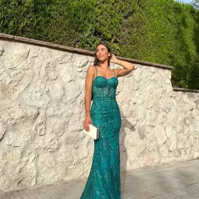 Vestido verde esmeralda brilhante de cerimónia com almaça fina e decote em coração, mala bege, em exterior junto a muro de pedra e sebes verdes