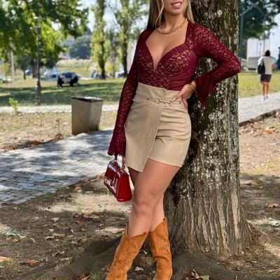 Mulher vestindo blusa de renda bordô, mini saia bege, botas castanhas e bolsa vermelha encostada numa árvore em parque