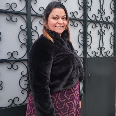 Mulher com casaco preto felpudo e saia roxa com padrão geométrico junto a portão de ferro decorativo negro