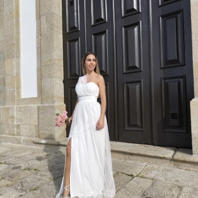 Mulher com vestido branco longo com fenda, segurando buquê de flores cor-de-rosa, em frente a porta de madeira escura.