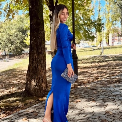 Mulher de vestido azul comprido com clutch brilhante e sandálias nude em ambiente exterior com árvores e calçada de pedra