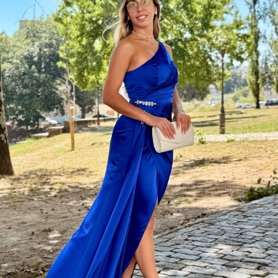 Mulher com vestido azul royal de um ombro só, sandálias brancas e mala bege ao ar livre