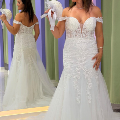 Vestido de noiva branco com renda e tule, decote coração, segurando bouquet branco, refletido em espelho num ambiente com arcos lilás e cortinas verdes.