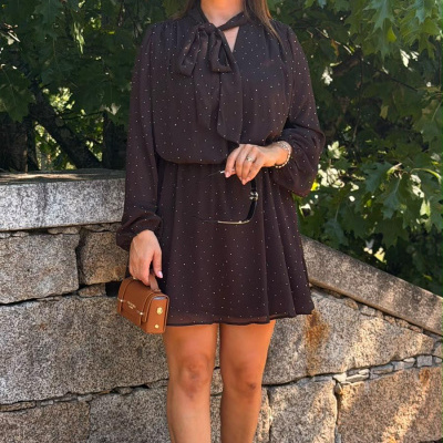 Vestido curto preto com padrão de pontos brancos, senhora com carteira castanha e sapatos cor de rosa junto a parede de pedra.