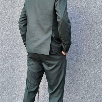 Homem de costas com fato verde-escuro e sapatos castanhos junto a parede cinzenta