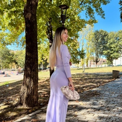 Mulher com vestido lilás, salto nude e mala estampada numa calçada de pedra sob árvores.