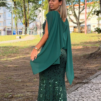 Mulher com vestido longo verde escuro com lantejoulas e capa fluida no parque