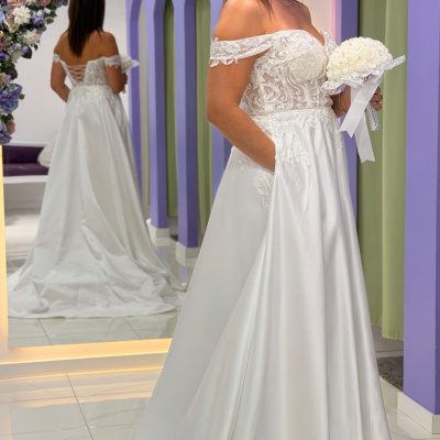 Mulher com vestido de noiva branco de renda e cetim, segurando ramo, em loja com decoração roxa e espelho.