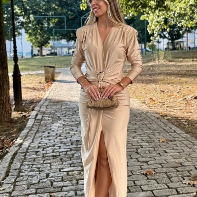 Mulher de vestido comprido bege com cinto dourado e sapatos castanho escuro, segurando bolsa dourada numa calçada ao ar livre.