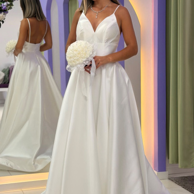 Vestido de noiva branco com alças finas e decote em V, mulher segura ramo branco, parede lilás e espelho floral ao fundo.